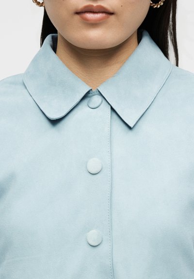 Artimos planas žmogaus, nešiojančio šviesiai mėlynę švarkelį su uždengtais mygtukais, ir auksinius vyniotus žiedinius auskarus.