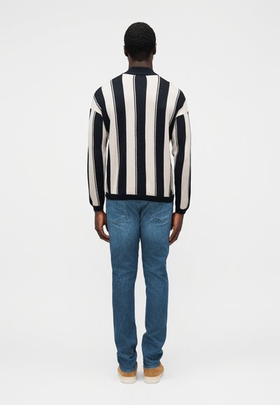 Ριγέ πουλόβερ σε κρεμ και ναυτικό μπλε, με στρογγυλή λαιμόκοψη και κατεβασμένους ώμους, συνδυασμένο με ανοιχτόχρωμα τζιν και μπεζ αθλητικά παπούτσια.