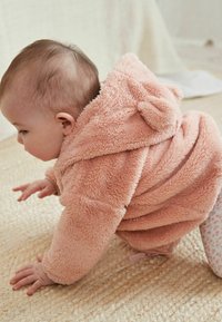 Enfant dans un manteau à capuche duveteux de couleur rose pastel avec des oreilles d'ours, rampant sur une surface tissée texturée, portant des leggings à motifs légers.