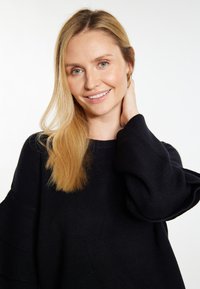 Musta ribbed neulepaita, jossa on leveät, röyhelöiset hihat. Mallilla on pitkät, suorat vaaleat hiukset ja hän koskettaa hellästi kaulaansa.