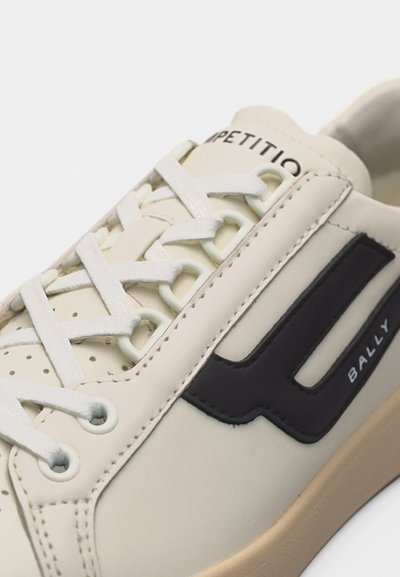 Gros plan d'une basket beige avec des lacets blancs, un patch logo noir sur le côté et le texte "COMPETITION" sur la languette.