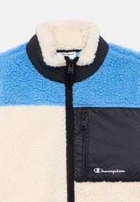 Casaco de fleece com painéis azuis e creme, bolso de peito com zíper preto, gola alta e logótipo da Champion no bolso e no puxador do zíper.