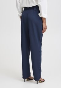 Pantalons adaptés bleu marine avec une coupe droite, présentant une texture lisse et une taille haute, associés à des sandales à talons noires.
