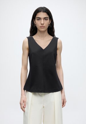 Vrouw met lang donker haar draagt mouwloze zwarte V-hals top en witte brede broek, staand tegen een effen witte achtergrond.
