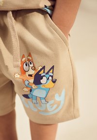 Shorts beiges à texture douce présentant un motif imprimé de deux chiens de dessin animé, un orange et un bleu, avec le nom "Bluey" en bleu clair.