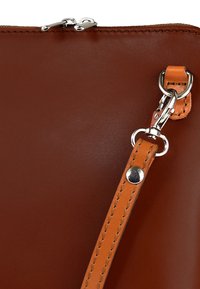 Bruine leren tas met een gladde textuur, een zilveren ritssluiting en een oranje afneembare band met metalen beslag en stikseldetail.