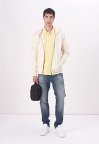 Licht crèmekleurige zip-up hoodie met oranje accenten, gele poloshirt, blauwe spijkerbroek en witte sneakers. Houdt een kleine zwarte tas vast.