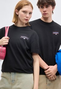 Δύο νέοι ενήλικες φορούν μαύρα μπλουζάκια με το λογότυπο Tommy· ο ένας κρατάει μια ροζ τσάντα, ο άλλος ένα μπλε μπουφάν, και οι δύο φορούν ανοιχτόχρωμο τζιν παντελόνι.