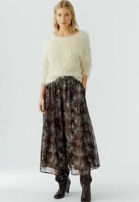 Flauschiger weißer Pullover mit Dreiviertelärmeln, kombiniert mit einem langen, dunklen Rock, der metallisch glänzende Blumenmuster aufweist, und kniehohen braunen Stiefeln.