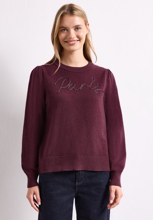 Femme souriante portant un pull violet avec des épaules bouffantes et "Paris" brodé sur la poitrine, associée à un jean bleu foncé.
