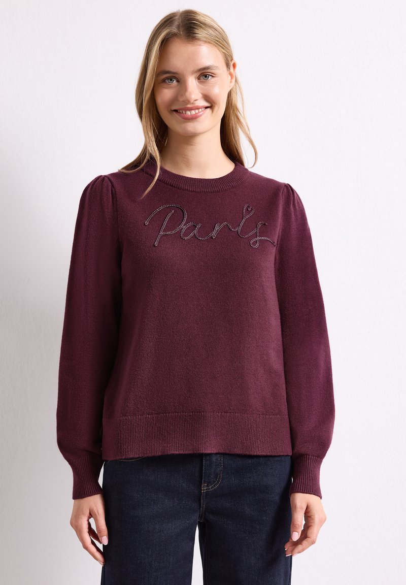Femme souriante portant un pull violet avec des épaules bouffantes et "Paris" brodé sur la poitrine, associée à un jean bleu foncé.