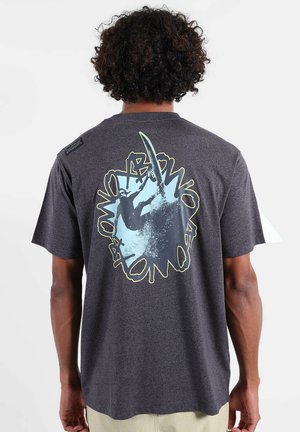 Homme aux cheveux bouclés portant un T-shirt gris foncé avec un motif de surfeur bleu et un texte circulaire au dos, debout devant un fond uni.