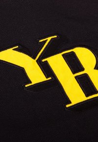 Zwarte stof met grote gele geborduurde letters "Y" en "B" met een gestructureerd oppervlak en contrasterende omranding.