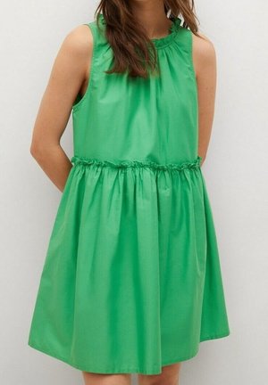 Femme portant une robe verte sans manches, longueur genoux, avec une taille froncée et un col à volants, sur un fond uni.
