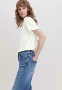 Valkoinen lyhythihainen paita yhdistettynä keskimittaisiin sinisiin farkkushortseihin. Kangas näyttää sileältä ja siinä on klassinen suora leikkaus sekä hienovaraiset vihjeet ompeluista.