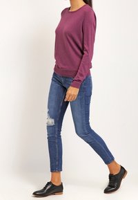 Fioletowy sweter z długim rękawem, okrągłym dekoltem, połączony z dopasowanymi niebieskimi dżinsami z przetarciami oraz czarnymi sznurowanymi butami na drewnianej podeszwie.