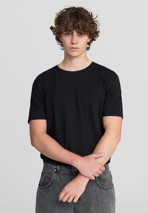 T-shirt nera a maniche corte con collo rotondo, realizzata in tessuto morbido, indossata con jeans in denim grigio dotati di tasche frontali e cuciture discrete.