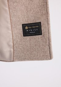 Beżowy płaszcz wełniany z teksturowanym, nakrapianym wzorem. Wyposażony w czarną metkę ze złotym napisem "Wool Collection" oraz "Makalu, wyprodukowano w Polsce."