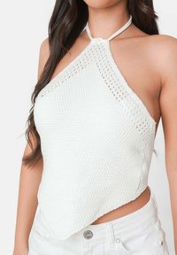 Witte gebreide haltertop met een textuurpatroon, driehoekige vorm en een centrale geknoopte band. Gecombineerd met witte high-waisted shorts.