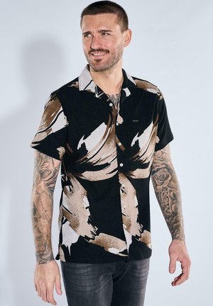 Mann mit kurzen Haaren und Tätowierungen an den Armen, trägt ein kurzärmeliges Hemd mit abstraktem Muster in Schwarz, Beige und Weiß und dunkle Jeans.