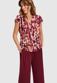 Kobieta w różowej bluzce z krótkim rękawem w ciemnoczerwone kwiatowe wzory oraz luźnych, wysokich, ciemnoczerwonych spodniach, z rękami w kieszeniach.