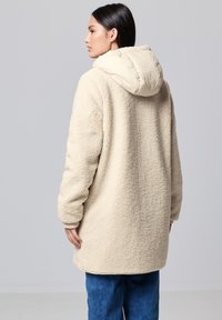 Mujer de perfil que lleva un abrigo de sherpa color crema con capucha acolchada y jeans azules, contra un fondo gris liso.