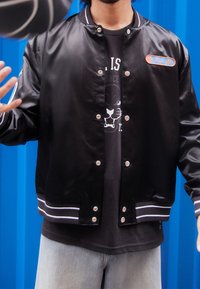 Black bomber jacket with a glossy finish, snap buttons, white striped cuffs, featuring a colorful patch. Worn over a dark graphic t-shirt.