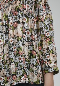 Blusa floreale con sfondo nero e fiori rosa, bianchi e verdi. Tessuto strutturato con maniche arricciate e dettagli con bottoni.