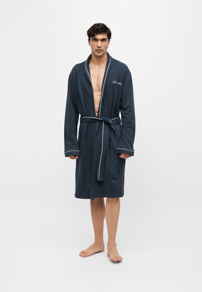 Homme debout pieds nus portant un peignoir bleu foncé jusqu'aux genoux avec un liseré blanc et une ceinture à la taille sur un fond clair uni.