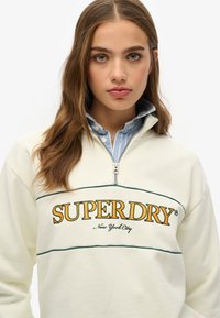 Crème kleurige zip-up sweatshirt met geborduurd "SUPERDRY" logo in oranje en groen, met daaronder een gekraagde blauw-wit gestreepte blouse.