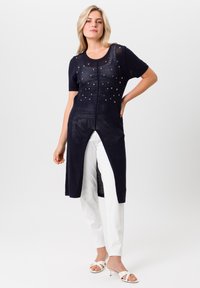 Marine gebreide lange cardigan met korte mouwen, featuring open-gehaakte patronen, decoratieve studs en zijsplitsingen, gecombineerd met witte broek.