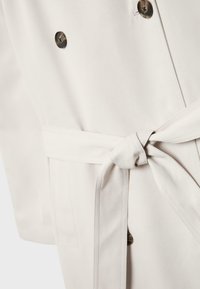 Nærbillede af en beige jakke med bindebælte i taljen og tre mørke knapper langs frontstolpen.