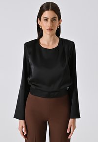 Blusa nera in satin a maniche lunghe con scollatura rotonda e dettagli arricciati sulle spalle, abbinata a pantaloni marroni aderenti. Texture liscia.
