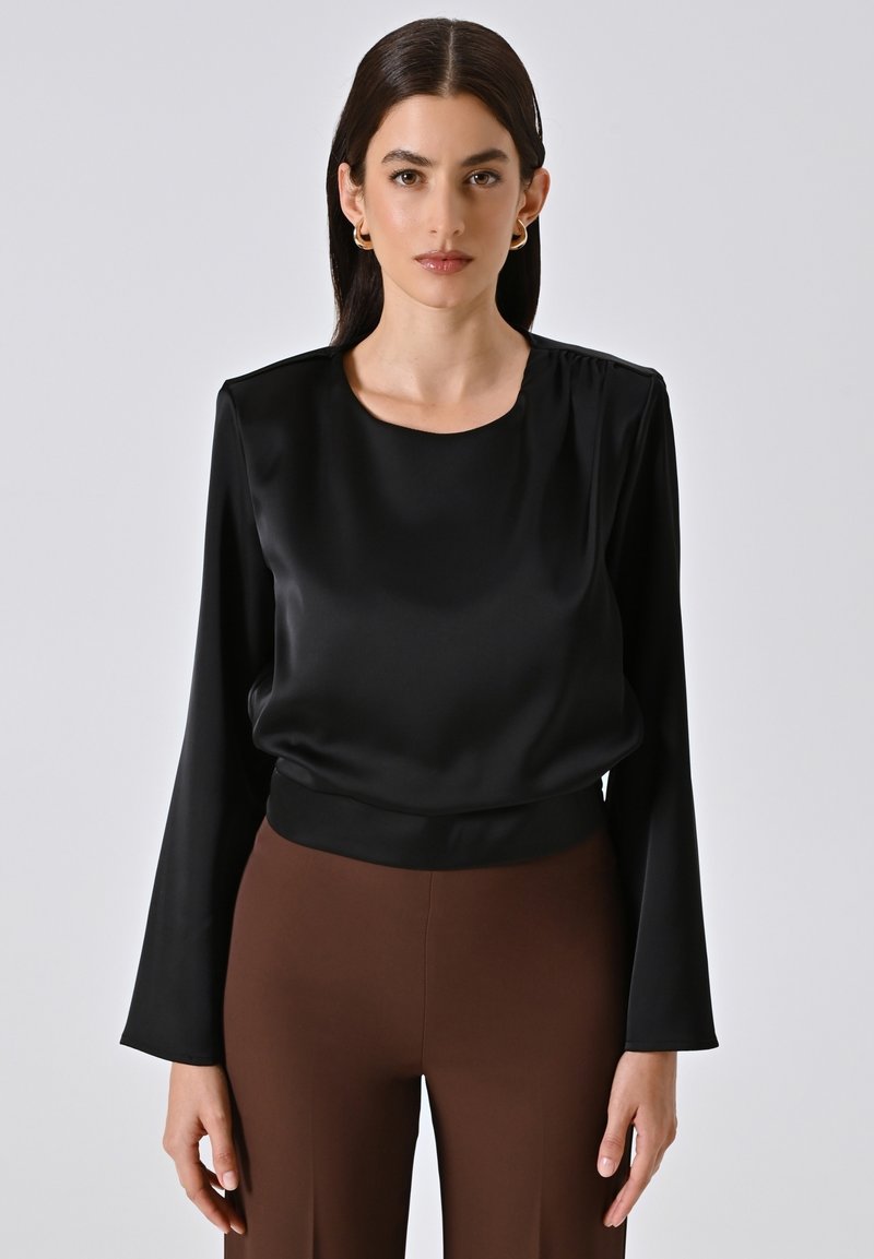 Blusa nera in satin a maniche lunghe con scollatura rotonda e dettagli arricciati sulle spalle, abbinata a pantaloni marroni aderenti. Texture liscia.