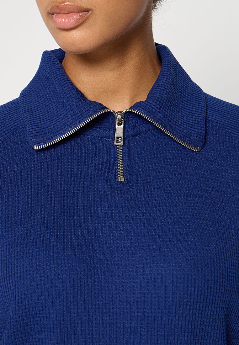 Niebieski sweter na zamek z teksturowanej, prążkowanej tkaniny. Posiada szeroki kołnierz i metalowy zamek, co sprawia, że ma zgrabny, a jednocześnie casualowy fason.