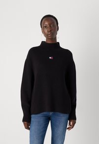 Svart strikkegenser med høy krage, prydet med en liten logo i rødt, hvitt og blått. Båret med lyseblå jeans, poserer modellen mot en hvit bakgrunn.