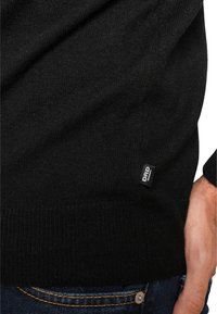 Black sweater in a fine knit texture with ribbed cuffs and hem. Features a small black label with "DRD" on the side.