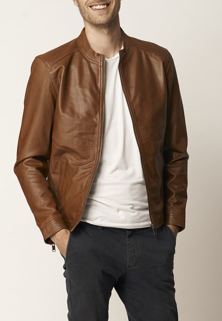 Giacca di pelle marrone con una texture liscia, chiusura con zip, colletto a coste e tasche laterali, indossata sopra una maglietta bianca e pantaloni scuri.