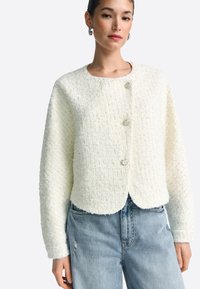 Balta tvīda jaka ar apaļu izgriezumu, trīs pogām priekšpusē, īsais piegriezums un viegli uzpūstām piedurknēm, kopā ar izbalējušām džinsa biksēm.