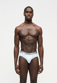 Izmos fekete férfi, szembenézve, fehér Calvin Klein alsónadrágot viselve, egyszerű világos háttér előtt.