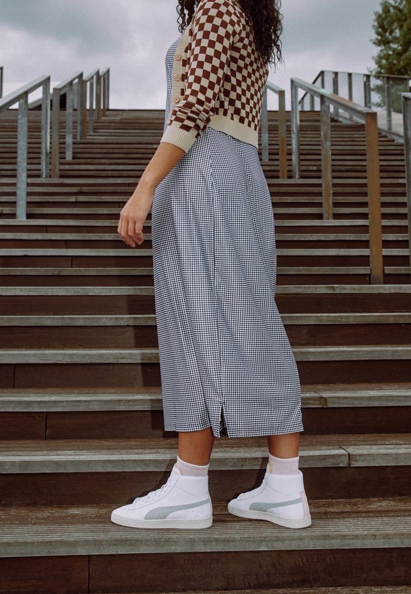 Personne debout de profil sur des escaliers en bois extérieurs, portant un pull à carreaux brun et beige, une longue jupe à motifs, et des baskets blanches.