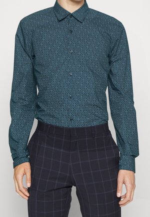 Chemise à manches longues en vert foncé avec un motif géométrique répétitif en bleu clair, de style boutonné et avec un col pointu. Associée à un pantalon à carreaux.