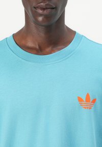 Teal bombažna majica s kratkimi rokavi in rebrastim ovratnikom, z majhnim oranžnim logotipom Adidas trefoil na levem prsnem predelu. Gladka tekstura.