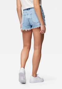 Vaaleansiniset farkkushortsit, joissa on rispaantunut helma ja kaksi takataskua, yhdistettyinä valkoisiin tennareihin. Kangas näyttää pehmeältä ja hieman haalistuneelta.