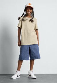 Camiseta de algodón beige con un pequeño logo, combinada con pantalones cortos de mezclilla azules y zapatillas altas blancas con un swoosh negro.