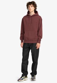 Bordeaux hættetrøje med kængurulomme, snoretræk og logo detalje på brystet, kombineret med mørke jeans og multicolore sneakers.