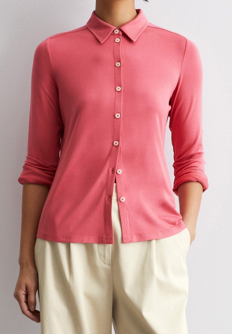 Personne portant une chemise rose à manches longues boutonnée, avec les manches retroussées et un pantalon beige clair, debout devant un fond uni.