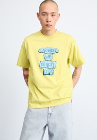 Kollane puuvillane T-särk, millel on sinine graafiline print, mis ütleb "CARHARTT WIP DETROIT U.S.A." Tavaline lõige lühikeste varrukatega.
