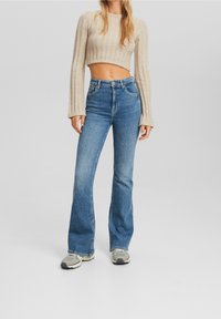 Woman wearing high-waisted flare blue jeans, beige cropped ribbed sweater, and gray sneakers standing against a plain gray background.