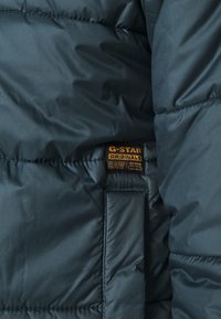 Ciemnozielona, ocieplana kurtka z naszywką z logo G-Star w kolorze złotym na rękawie. Gładka faktura z pikowanymi detalami przeszyć.